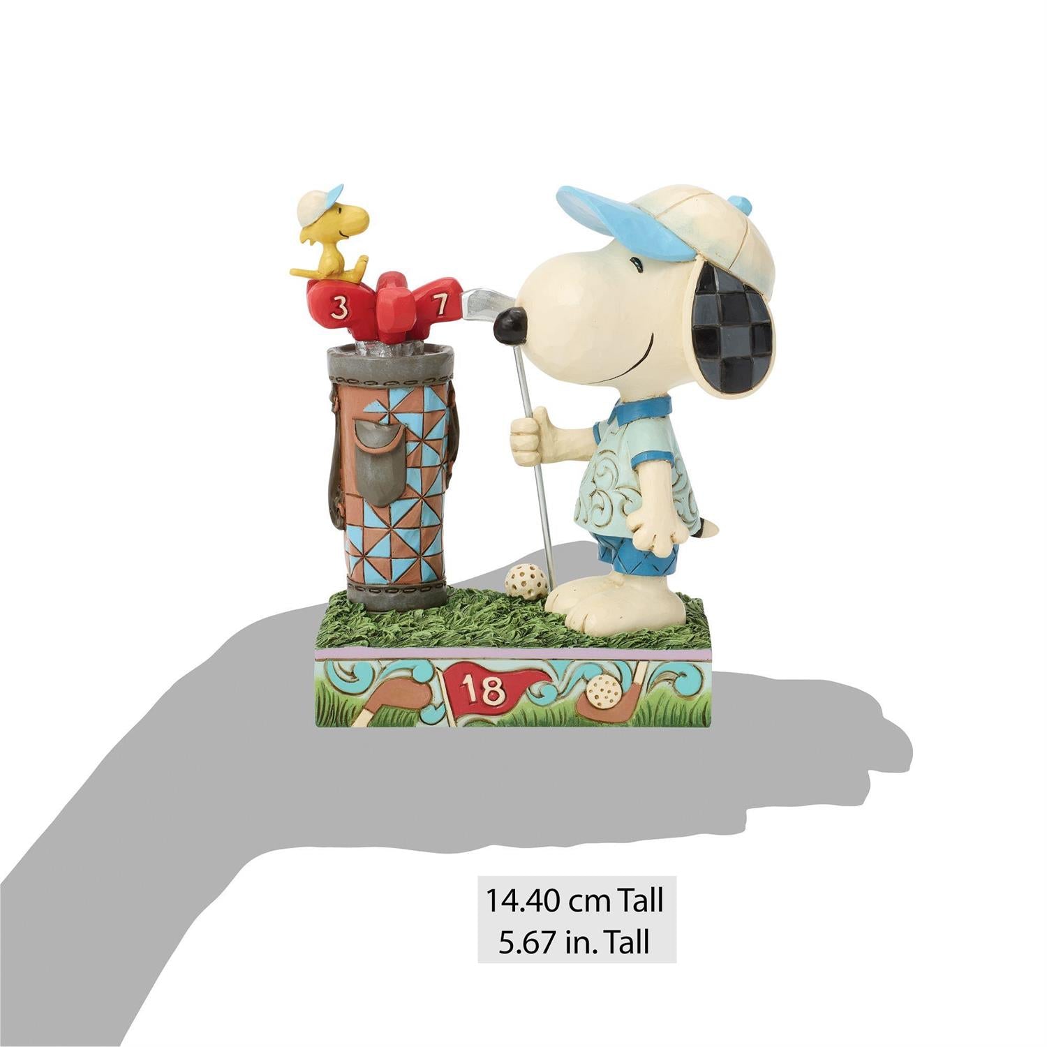 Snoopy dressed in his Golf clothes holding a Golf Club standing next to a Golf bag while Woodstock sits on the Golf Clubs and watches